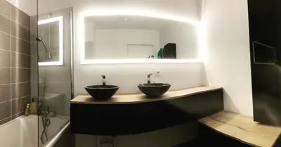 Salle de bain avec deux vasques posées sur un plan sombre, un miroir lumineux et une baignoire